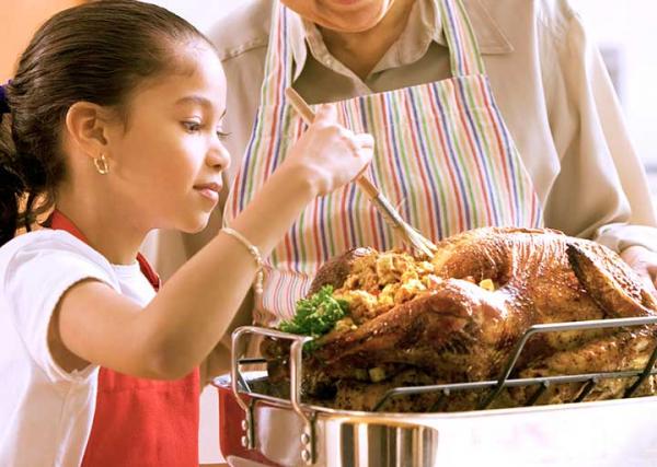 A young girl bastes a turkey in the kitchen while her grandmother watches.