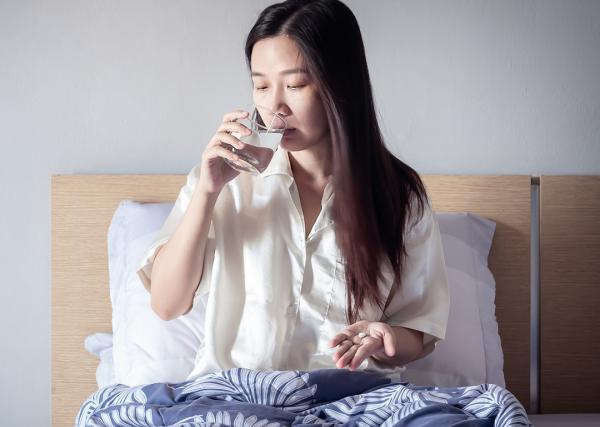 A young woman takes a pill with a glass of water from her bed.