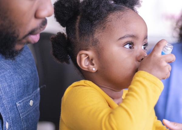 A young child sitting on her father’s lap uses an inhaler with the help of a healthcare provider.