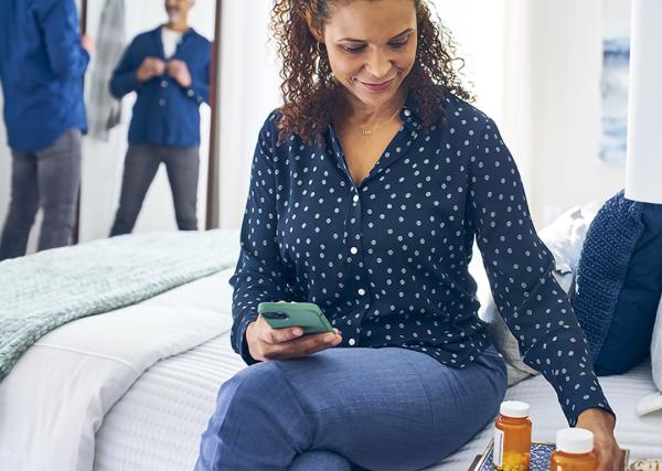 A woman sits on the edge of her bed and checks a prescription label on her nightstand.