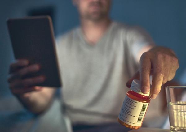 : A man sits up in bed to check a medication on his side table.