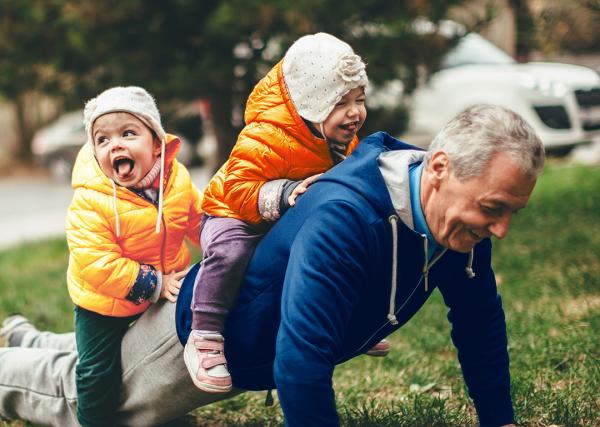 An older man does pushups outside with his two young grandchildren sitting on his back.