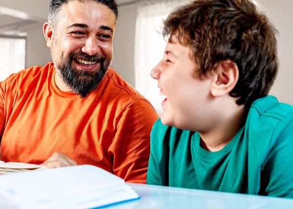 A father and his young son laugh and smile while the father helps his son with homework.