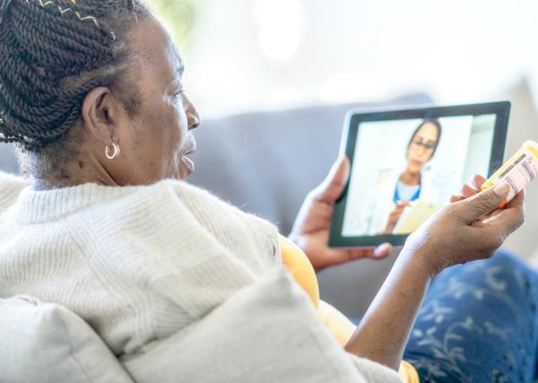 A woman holding a prescription medication bottle in one hand and a tablet in the other talks to a doctor through a telehealth visit.