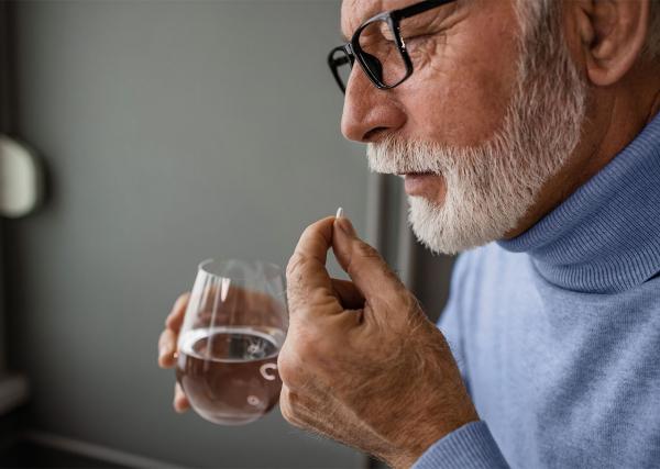 An older man holds a medication tablet in one hand and a glass of water in the other.