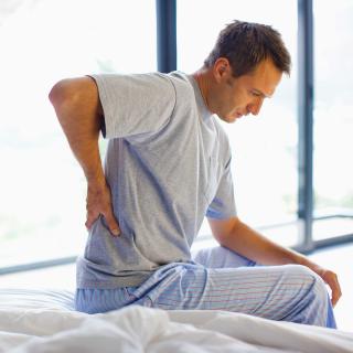 A man sits on the edge of his bed and rubs his lower back in pain.