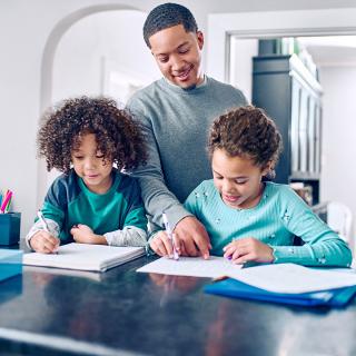 A father helps his two young children with their homework at the kitchen counter.