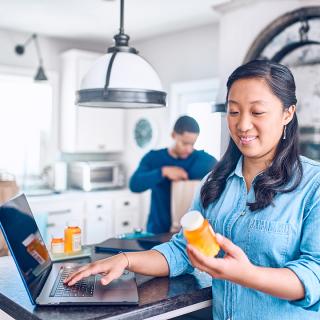 A woman checks the label on her prescription pill bottle in her kitchen while her husband unloads groceries in the background.