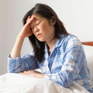 A woman suffering from a migraine sits up in bed while closing her eyes and holding her head.