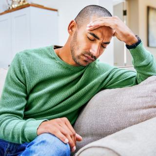 A man with a headache sits on his couch with his eyes closed, holding a hand to his head.