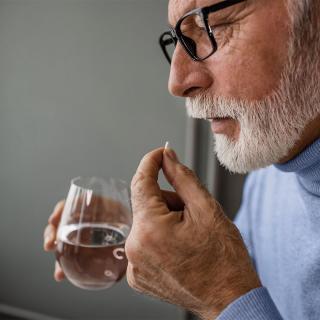 An older man holds a medication tablet in one hand and a glass of water in the other.